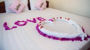 A bed with a white sheet, pink pillows, and a towel arranged in a heart shape with purple flower petals.