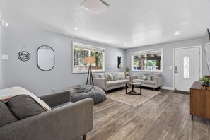 Ein modernes Wohnzimmer mit einem grauen Sofa, einem Couchtisch und Holzboden. Es gibt zwei Fenster mit Vorhängen, eine Tür und einen Spiegel an der Wand.