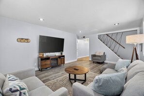 A modern living room with a flat-screen TV, a wooden entertainment unit, a grey sofa, a round coffee table, and a staircase with a railing. - Family-Friendly Tahoe Home w/ 2 Living Rooms, 3BR, Pack n Play, Near Heavenly Skiing (South Lake Tahoe)