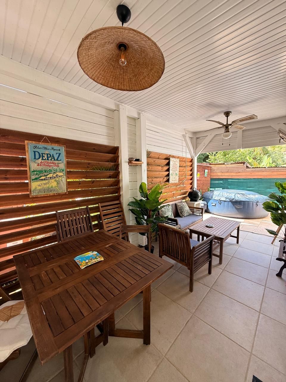 A covered outdoor seating area with a wooden table and chairs, a hanging wicker lamp, and a sign on the wall.