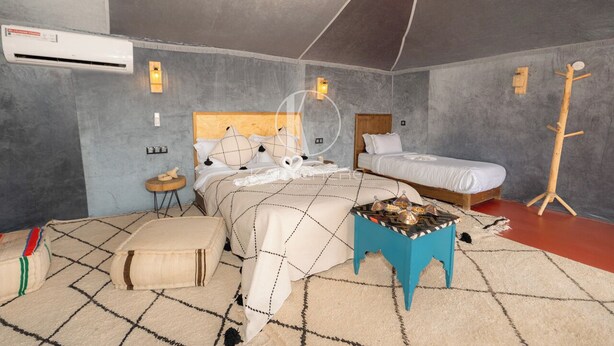 Un dormitorio con cama, mesitas de noche, un taburete pequeño y un equipo de aire acondicionado.
