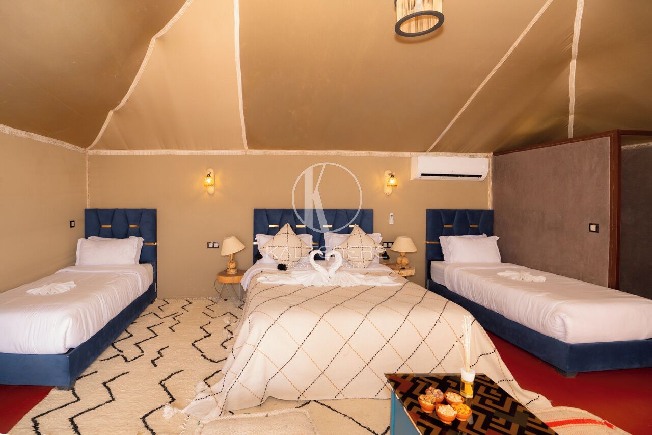 A room with two beds, a large bed with a decorative headboard, and a smaller bed. There are wall-mounted lights and an air conditioning unit.