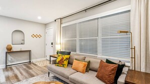 A living room with a sofa, a coffee table, a lamp, and a window with blinds. - September Savings:15% Off All 3+ Night Stays! (Charlotte)