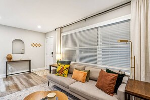 A living room with a sofa, a coffee table, a lamp, and a window with blinds. - September Savings:15% Off All 3+ Night Stays! (Charlotte)