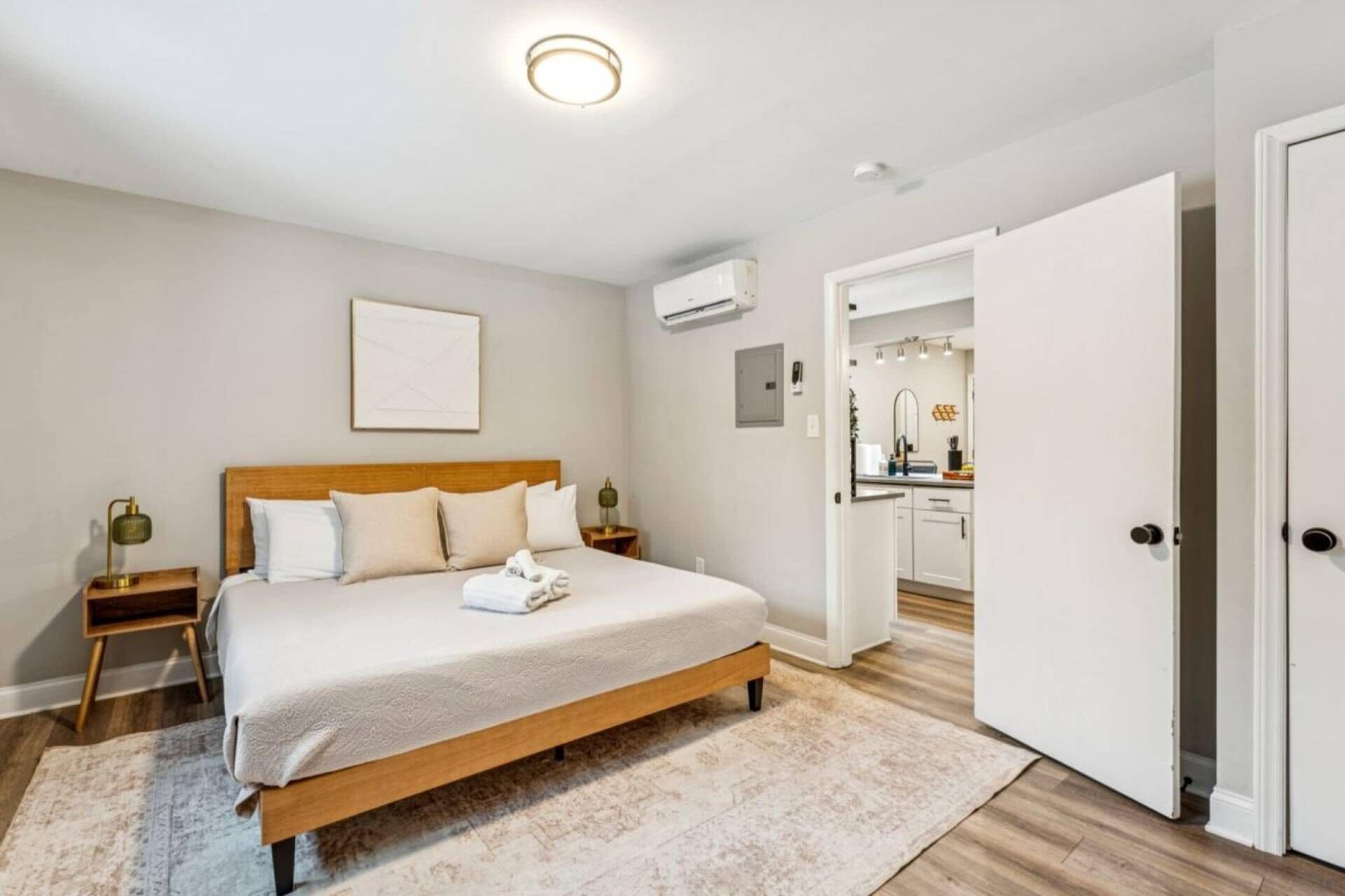 Une chambre avec un lit en bois, une table de chevet avec une lampe, un climatiseur fixé au mur et une porte donnant sur une salle de bain.