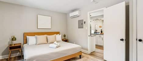 Une chambre avec un lit en bois, une table de chevet avec une lampe, un climatiseur fixé au mur et une porte donnant sur une salle de bain.