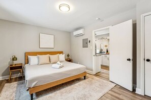 A bedroom with a wooden bed, a bedside table with a lamp, a wall-mounted air conditioner, and a door leading to a bathroom. - Modern & Cozy Oasis In Noda | Community Gym, Dog Park & Courtyard with Firepit | Pets OK! +20% Off! (Charlotte)