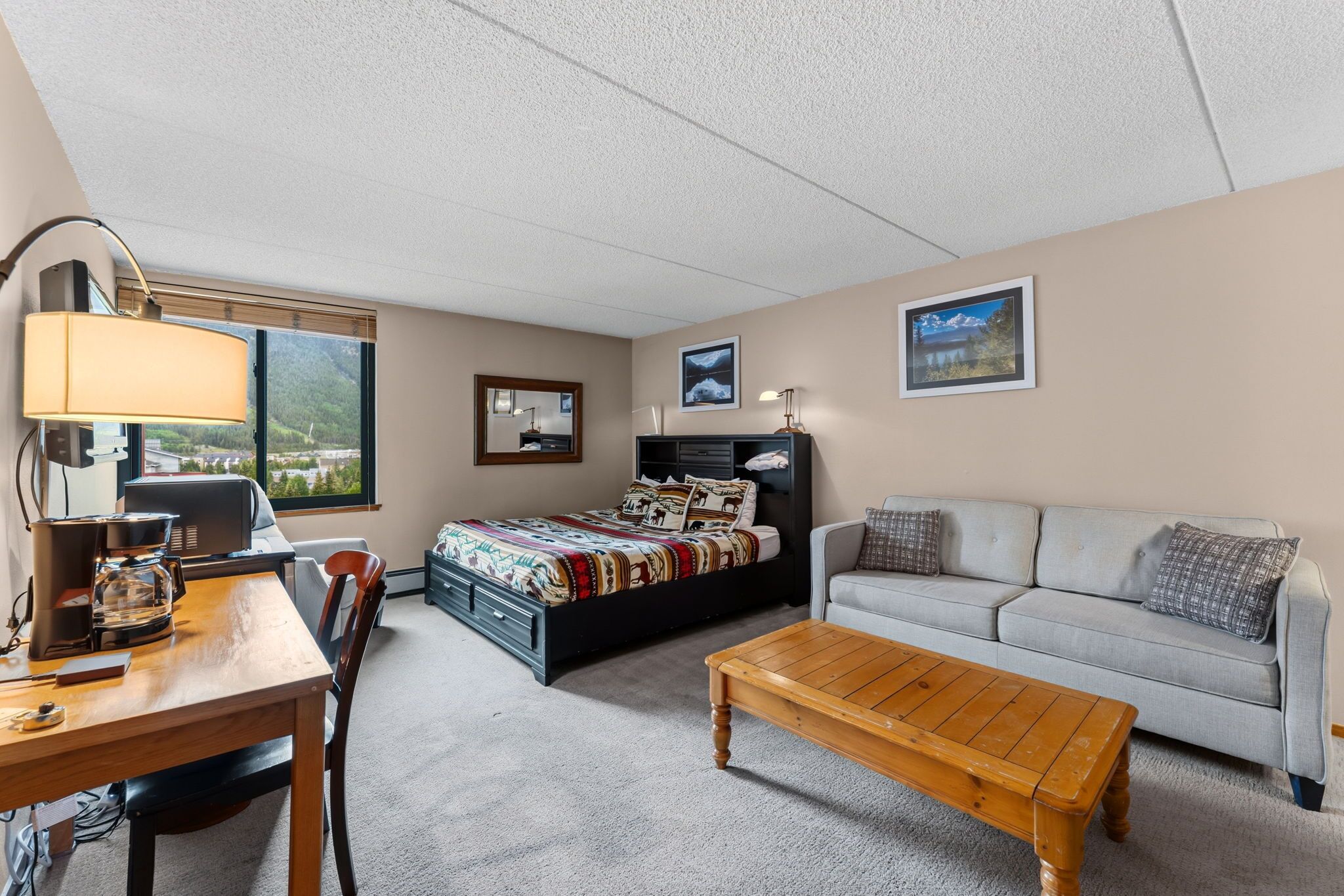 Habitación de hotel con cama, sofá, mesa de centro, escritorio con lámpara y vista a la montaña desde la ventana.