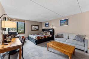 A hotel room with a bed, a sofa, a coffee table, a desk with a lamp, and a view of a mountain through the window. - Copper Mountain Studio in Center Village Walk to Lifts and Dining (Frisco)