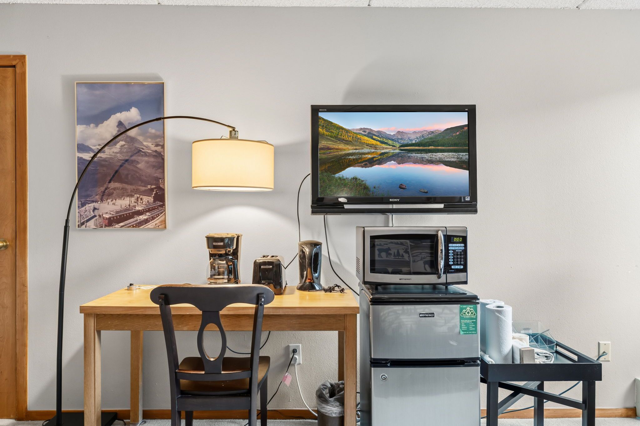 Una habitación con escritorio de madera, una silla, televisor montado en la pared, lámpara, cafetera, tostadora, microondas y refrigerador.