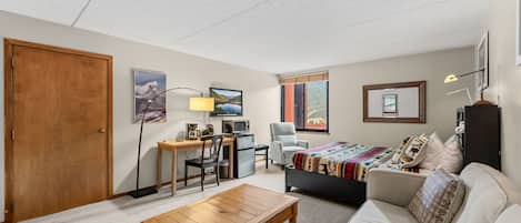Un salon meublé comprenant une table basse en bois, un canapé, un bureau avec une chaise, un téléviseur et un lit recouvert d’une couverture colorée.