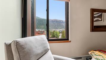 Una habitación con ventana que da a un paisaje montañoso, una silla y una cama con colcha estampada.