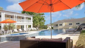Un área de piscina con sillones de descanso, una sombrilla y un edificio de dos pisos con balcones.