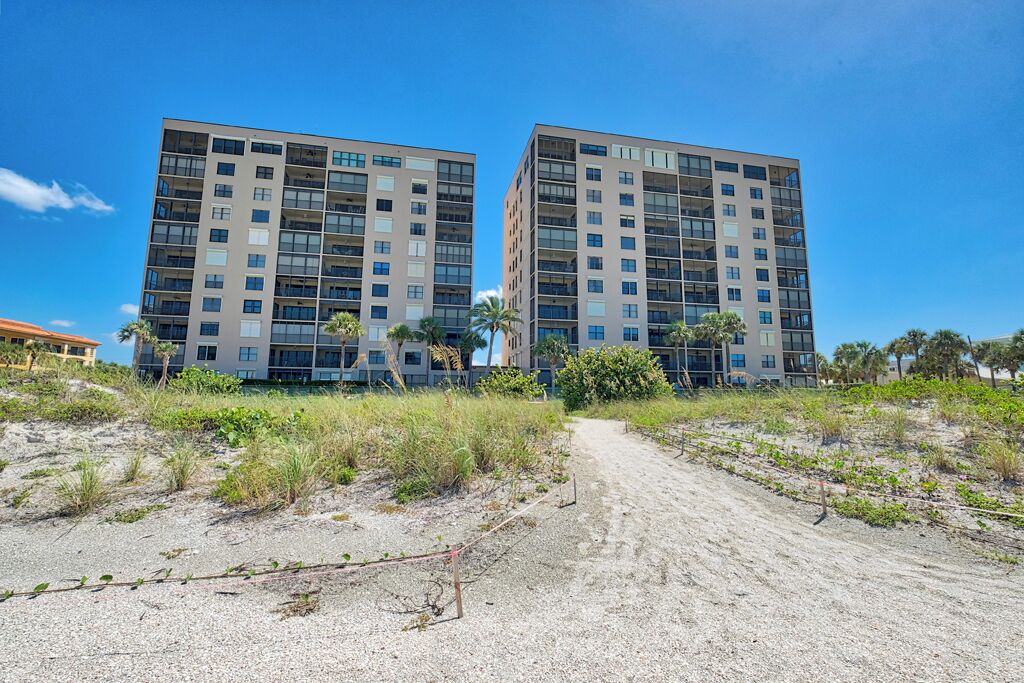 A modern building with multiple floors and balconies, surrounded by a sandy area with sparse vegetation.