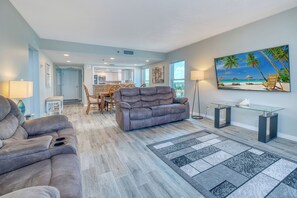 A modern living room with a grey sofa, a dining area with wooden chairs, and a kitchen in the background. - Direct Gulf Front Corner Unit with Expansive Views (Indian Rocks Beach)