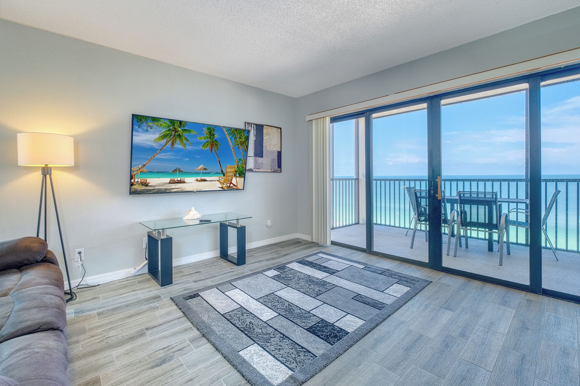 A modern living room with a beach-themed painting, a glass coffee table, a sofa, and a balcony with a view of the ocean.