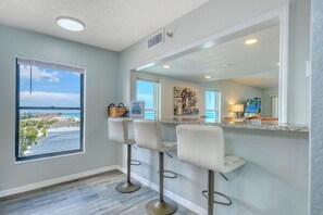 A kitchen with a large window, a bar area with stools, and a view of the ocean. - Direct Gulf Front Corner Unit with Expansive Views (Indian Rocks Beach)
