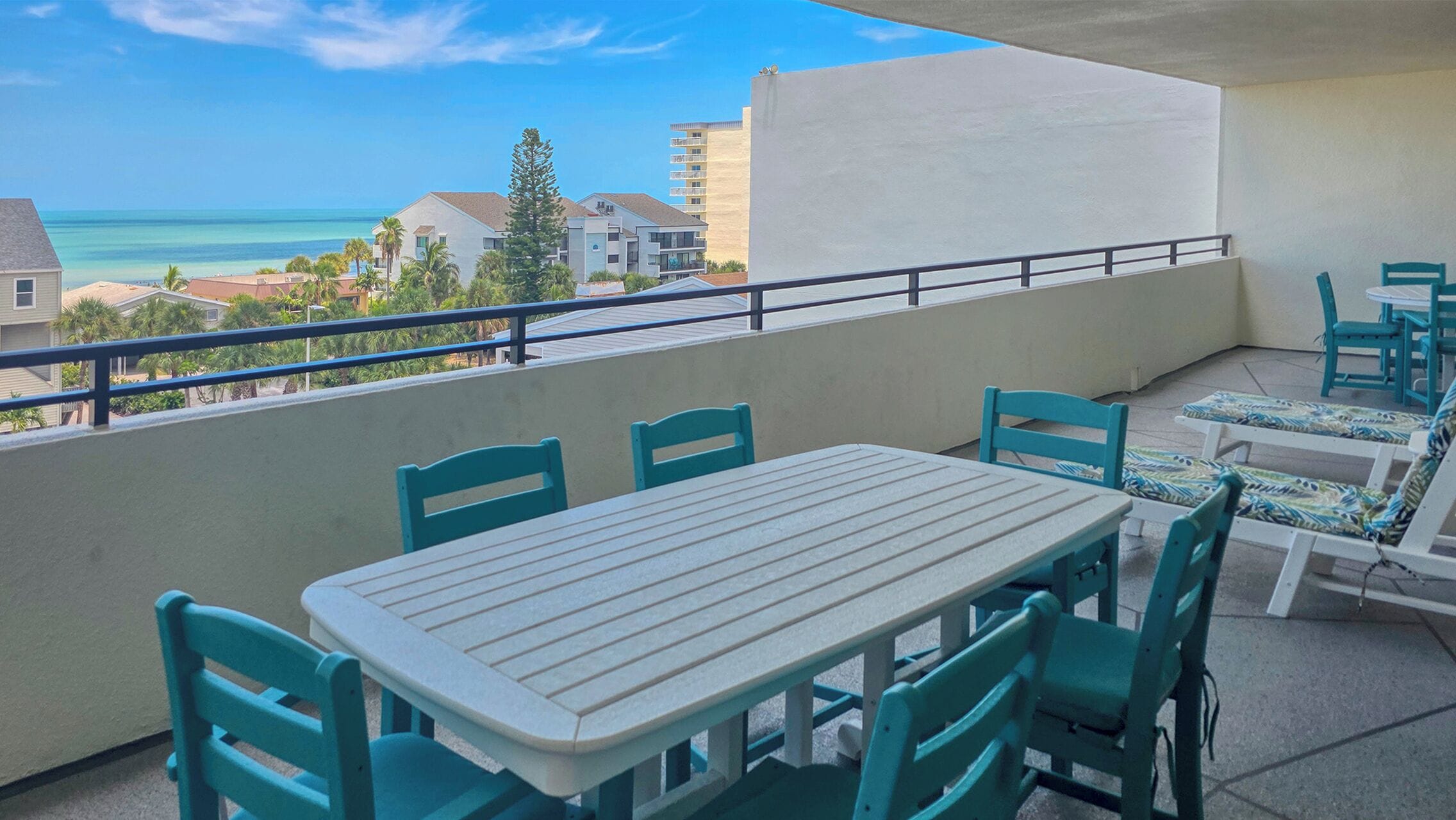 A balcony with a table and chairs, overlooking a coastal view with buildings and palm trees.