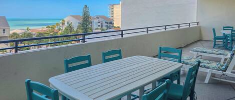 A balcony with a table and chairs, overlooking a coastal view with buildings and palm trees.