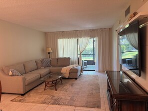 Ein Wohnzimmer mit einem Ecksofa, einem Glastisch, einem Flachbildfernseher und einer Schiebetür zu einem Außenbereich.