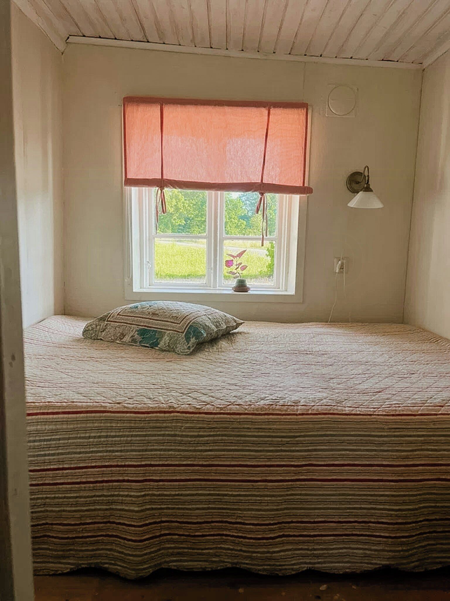 A small room with a single window, a bed with a striped bedspread, and a wall-mounted lamp.