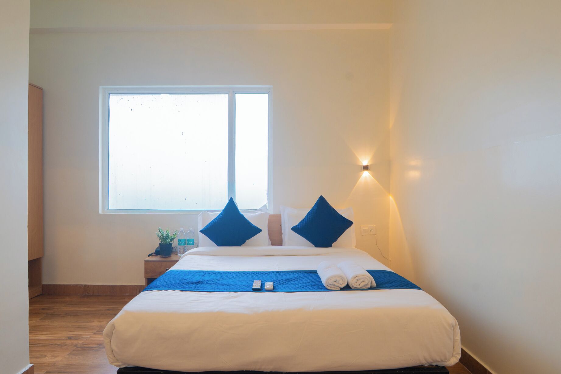 A hotel room with a bed, pillows, a bedside table with a plant and water bottles, a window, and a wall-mounted light.
