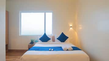 A hotel room with a bed, pillows, a bedside table with a plant and water bottles, a window, and a wall-mounted light.