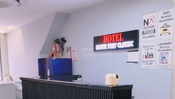 Un comptoir de réception d’hôtel avec une enseigne indiquant « Hôtel Silver Stay Classic » et mentionnant les services suivants : « Service de taxi disponible », « Service de blanchisserie disponible » et « Il est interdit d’apporter de la nourriture de l’extérieur ».