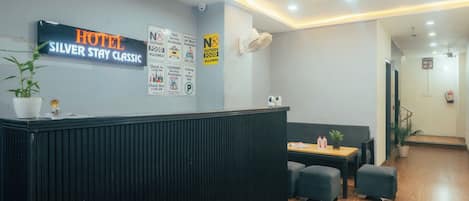 A hotel reception area with a sign reading 'HOTEL SILVER STAR CLASSIC,' a potted plant, a reception desk, a seating area with a table and chairs, and various posted signs indicating services and policies.