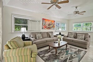 Ein Wohnzimmer mit zwei Sofas, einem Couchtisch und einem gestreiften Sessel. Es gibt Deckenventilatoren, ein Gemälde und Fenster mit Jalousien.