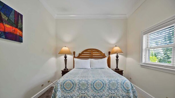 A bedroom with a bed, two bedside lamps, a window with blinds, a rug, and a wall painting. - Comfortable Villa Near Beach + Atlantis + Downtown (Nassau)