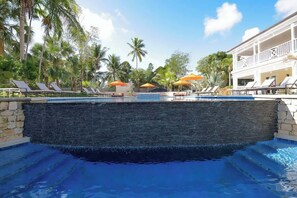 Ein Swimmingpool mit Liegestühlen und Sonnenschirmen, umgeben von Palmen und einem Gebäude mit einem Balkon.