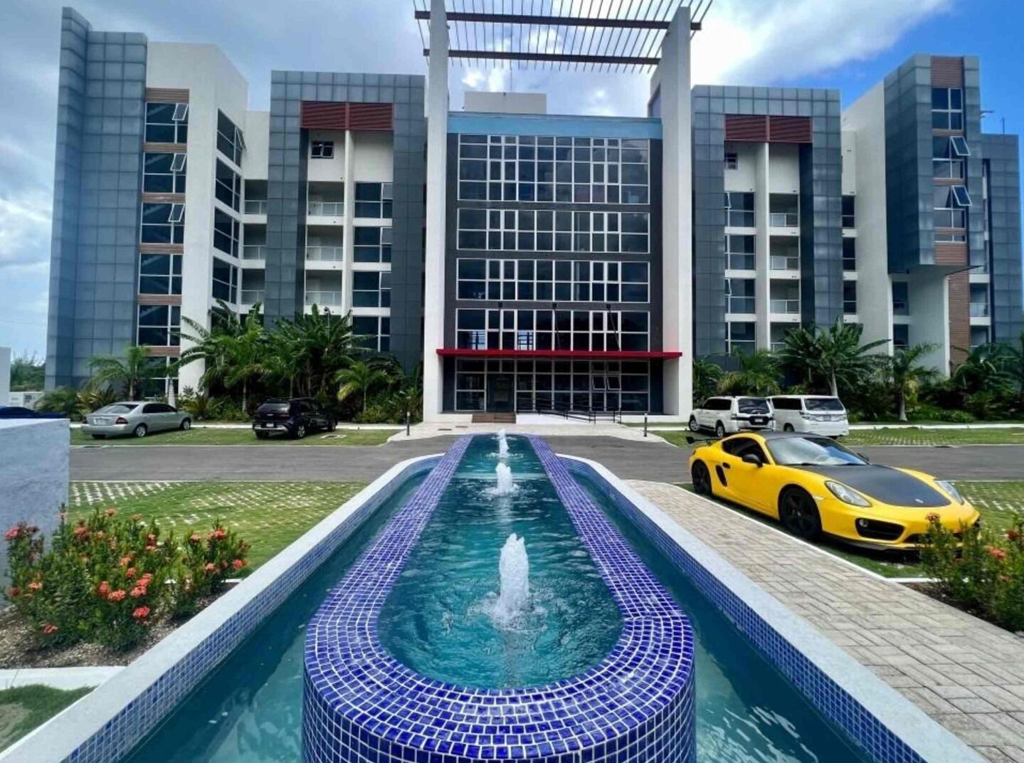 Ein modernes Gebäude mit einem Pool im Vordergrund, umgeben von parkenden Autos und Palmen.
