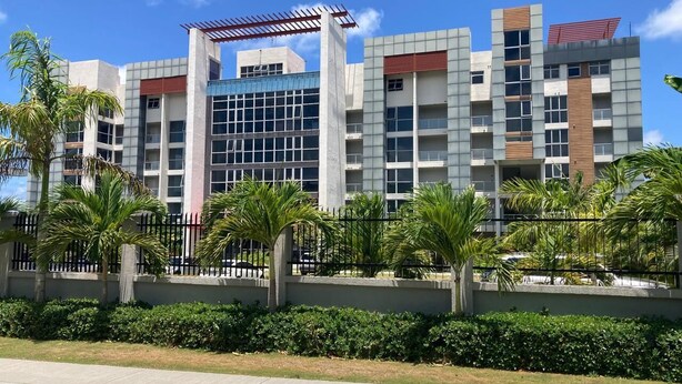 A modern multi-story building with a mix of glass and concrete facades, surrounded by a low hedge and palm trees. - 3BR Luxury Penthouse Suite - Dream 36  (Montego Bay)