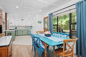 A dining area with a blue table and chairs, a kitchen in the background, and a view of trees through a window. - 3BR Oceanside with Easy Beach Access | Golf cart + Pet Friendly (Emerald Isle)