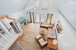 A spacious room with a wooden floor, a ceiling fan, a television, a sofa, a coffee table, and a small wooden cabinet. - 3BR Oceanside with Easy Beach Access | Golf cart + Pet Friendly (Emerald Isle)