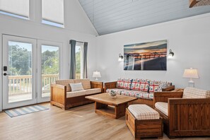 A spacious living room with a wooden floor, a sofa set, a coffee table, and a painting of a sunset over a pier. - 3BR Oceanside with Easy Beach Access | Golf cart + Pet Friendly (Emerald Isle)