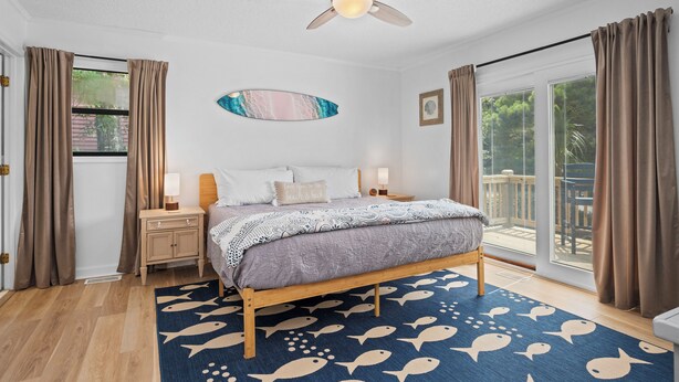 A bedroom with a bed, nightstands, a ceiling fan, and a view of the outdoors. - Pet-Friendly Cottage w/ Golf Cart, Kayak & Easy Beach Access (Emerald Isle)