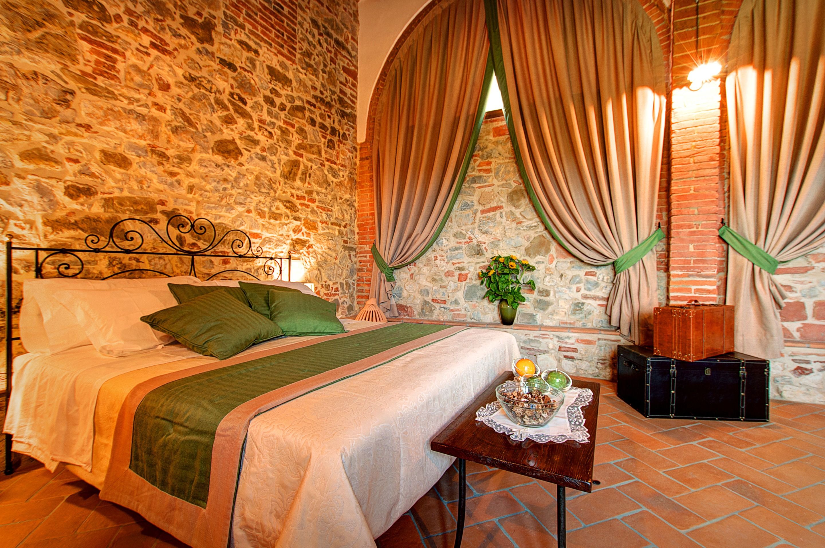 A bedroom with a stone wall, a bed with green pillows, a small table with a vase and fruit, and a suitcase.