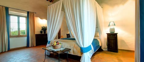 A bedroom with a four-poster bed, a bedside table with a fruit basket, a lamp, and a window with curtains.
