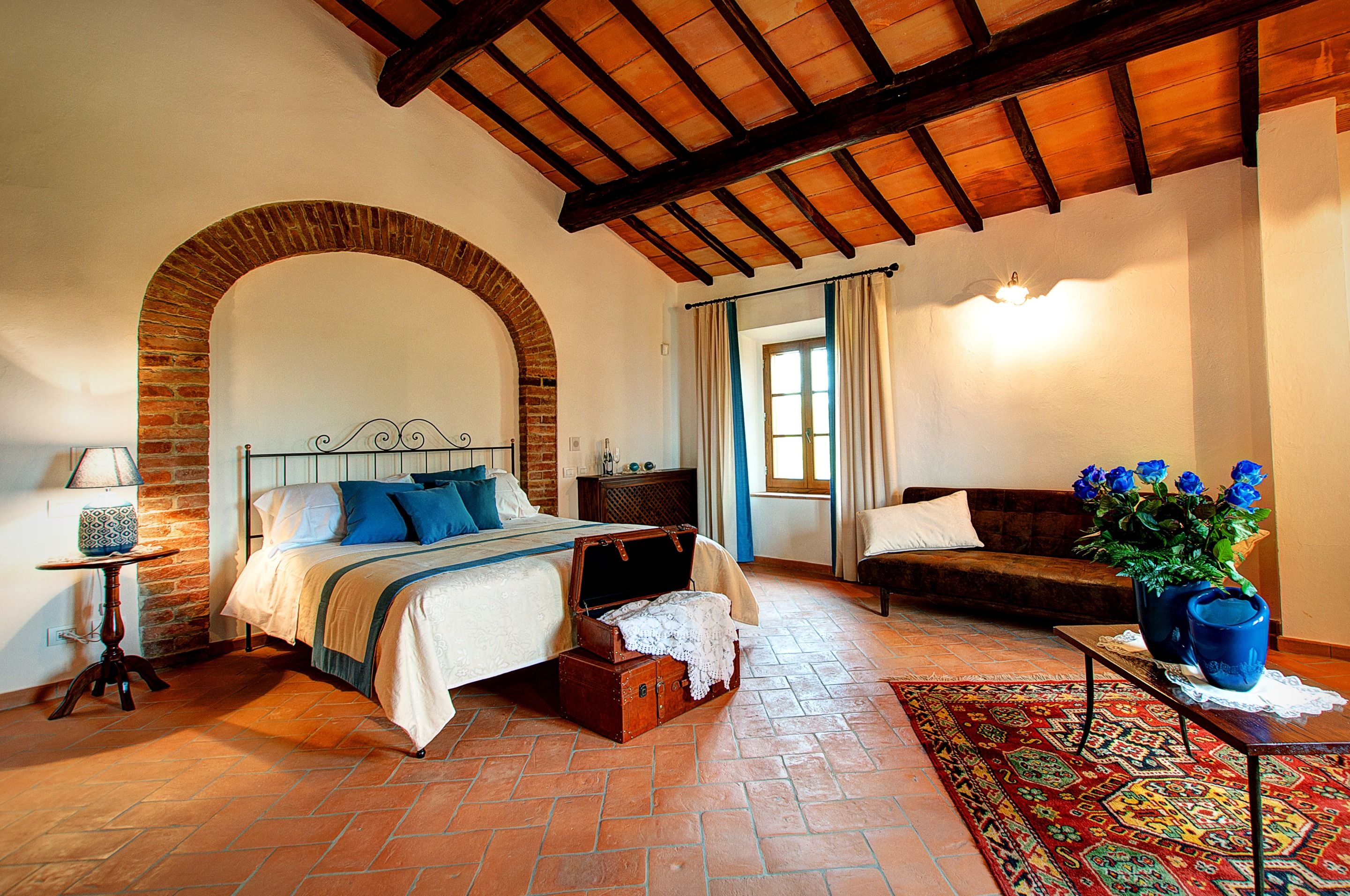 A bedroom with a large bed, a chest of drawers, a bench, a table with a vase of flowers, and a patterned rug.