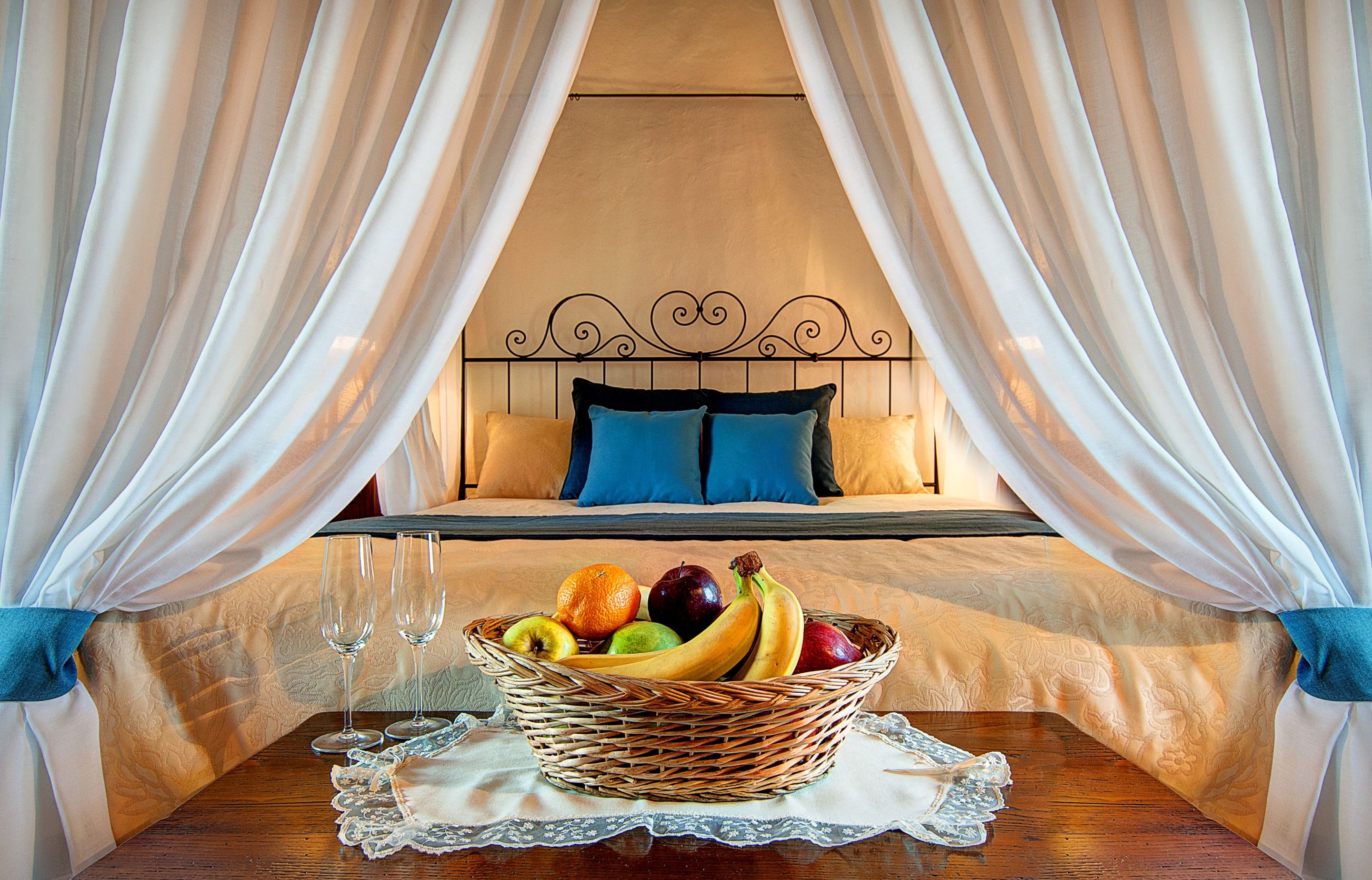 A hotel room with a bed, a basket of fruit, and two glasses on a table.