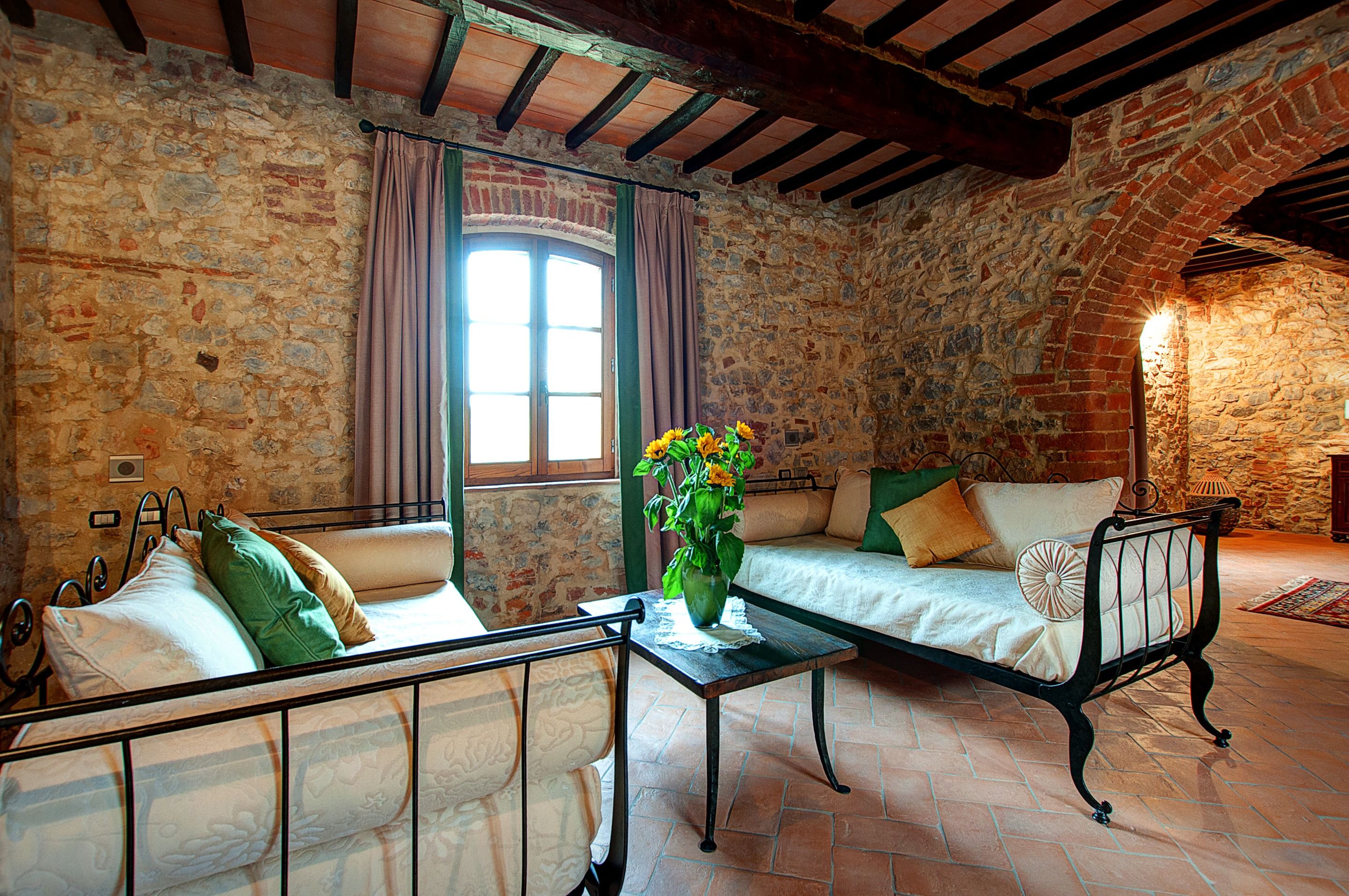 Una habitación rústica con paredes de piedra, techo de madera, cama con almohadas verdes y beige, sofá con funda blanca y una mesa con un jarrón de flores.