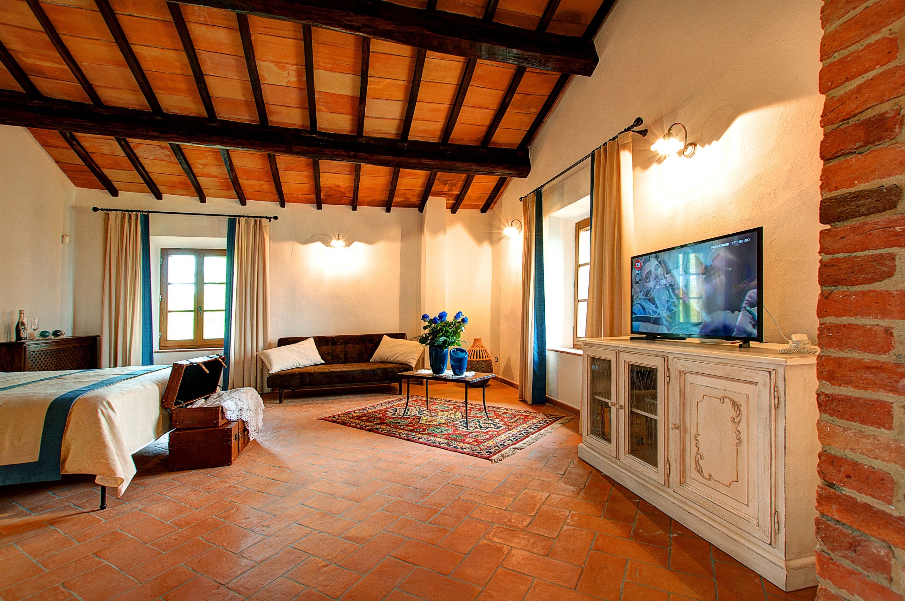 A cozy room with a wooden ceiling, a television on a cabinet, a sofa, a small table with a vase of flowers, and a bed with a suitcase.