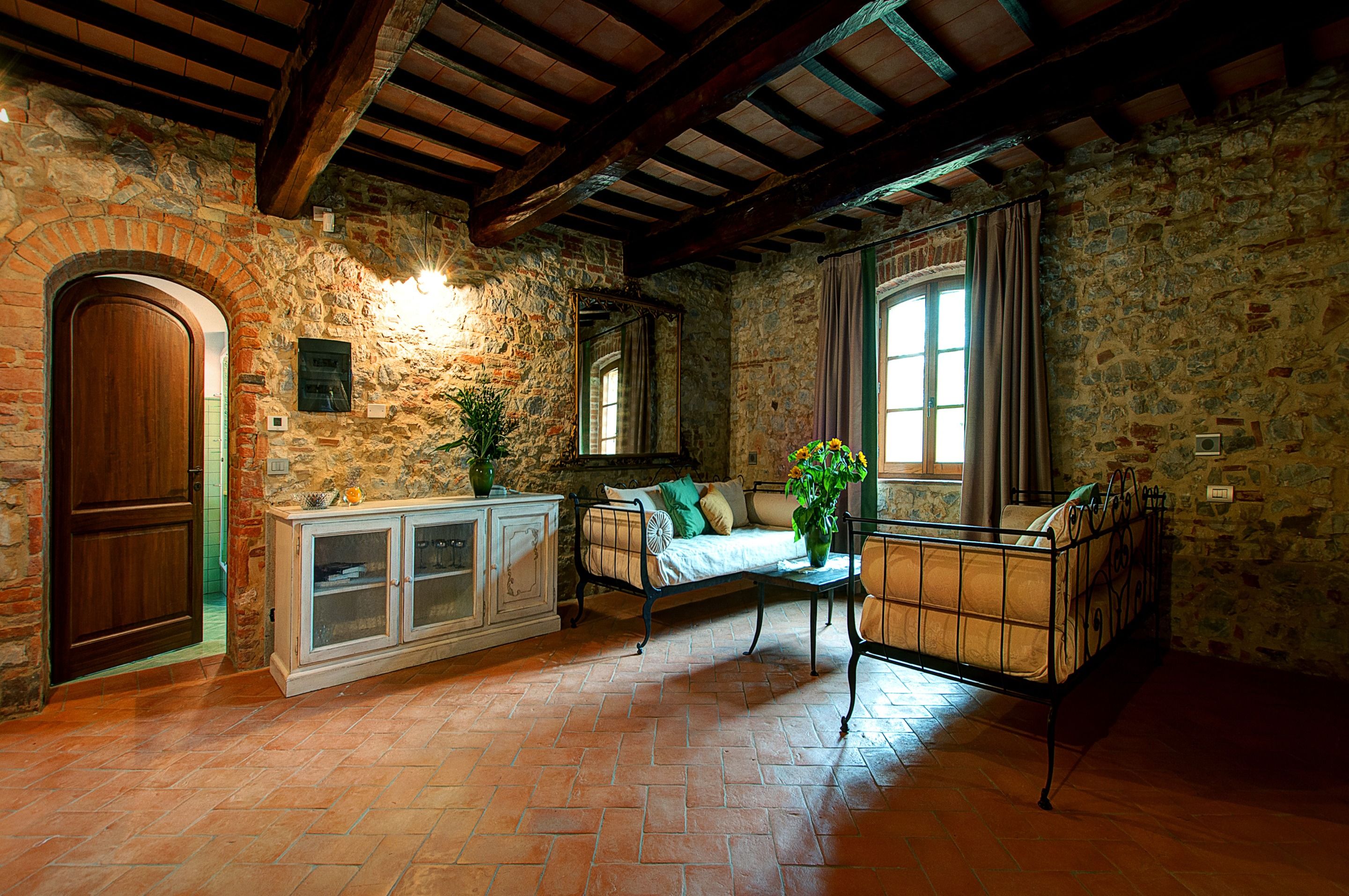 A rustic room with stone walls, a wooden door, a cabinet, a sofa, a table with a vase, and a window with curtains.
