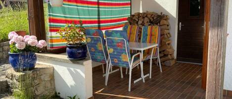Une terrasse avec une nappe à rayures, une table avec des chaises, une plante en pot et une pile de bois de chauffage.