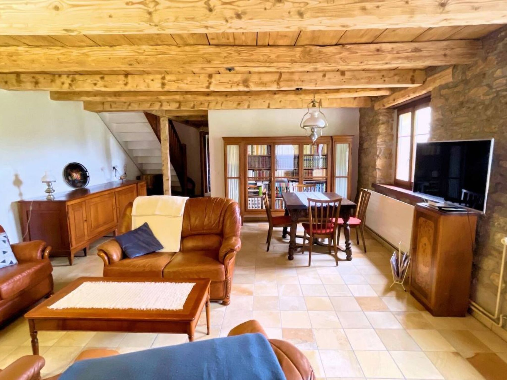 A spacious living area with a wooden ceiling, a dining table with chairs, a leather sofa, a television, and a bookshelf.