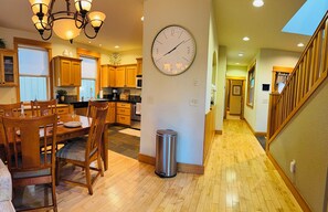 A spacious kitchen with wooden cabinets, a dining area with a wooden table and chairs, a wall clock, and a trash can.