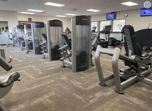 A gym with various exercise machines, a television screen, and a visible exit sign.