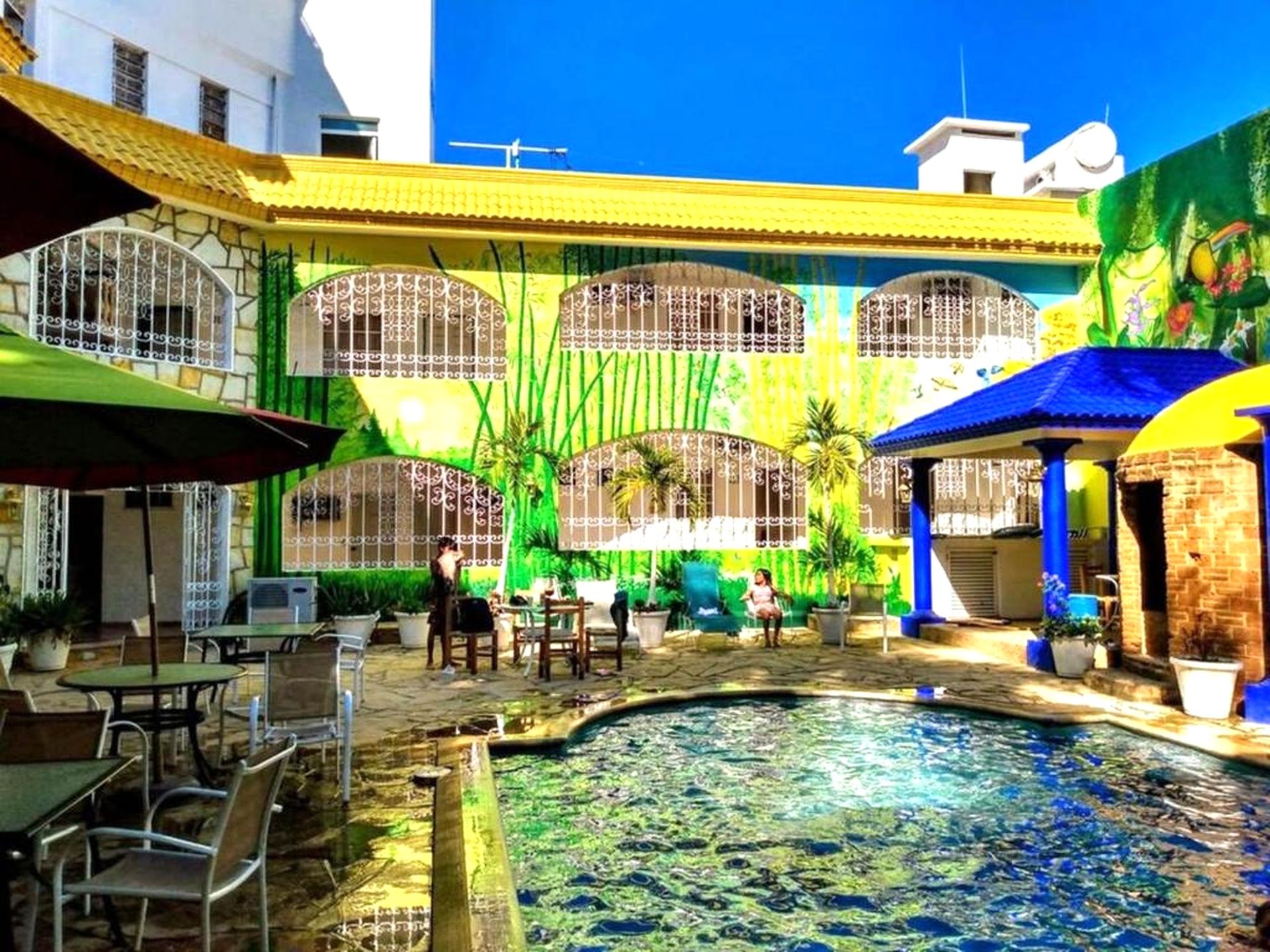 Un patio con piscina, mobiliario exterior y un edificio con grandes ventanales y un mural.